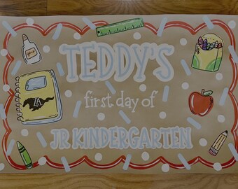 Puede incluir: Un letrero pintado a mano para "Teddy's first day of Jr Kindergarten". El letrero presenta ilustraciones de útiles escolares como un cuaderno, crayones, pegamento, una regla, una manzana y un lápiz. El letrero está decorado con un borde rojo y blanco.