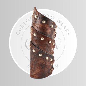 May include: Brown leather arm cuff with gold-colored studs. The cuff is made of multiple layers and has a distressed finish. The text "CUSTOM TO WEARS" is visible in the background.