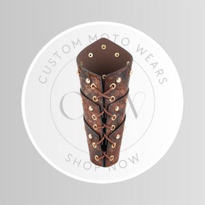 May include: Brown leather arm guard with a textured pattern, laced with brown cord and secured with gold-colored rivets. The arm guard is designed with a pointed top and is likely for protection or fashion. The image includes the text "CUSTOM MOTO WEARS SHOP NOW".