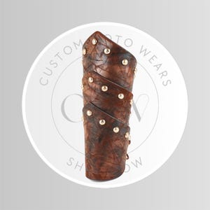 May include: Brown leather arm guard with gold-colored studs. The arm guard is made of layered leather and has a textured appearance. The product is designed to protect the forearm.