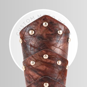 May include: Close-up of a brown leather arm guard with gold-colored studs. The leather has a textured pattern and a distressed appearance. The text "CUSTOM MOTO WEAR" is visible in the background.