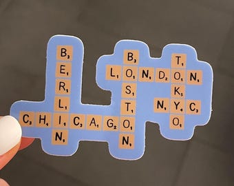 May include: A light blue magnet with the names of cities spelled out using Scrabble tiles. The cities include Berlin, Chicago, London, and New York City. The magnet is held by a hand with white nail polish.