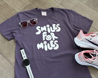 May include: A purple t-shirt with the text "Smiles for Miles" in white with a smiley face graphic. The shirt is laying on a wood floor with a white watch and pink running shoes.