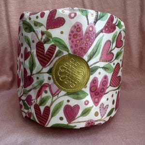 May include: A white cylinder wrapped in paper with a pink and green floral pattern featuring hearts and gold dots. A gold seal with a knot design is in the centre of the cylinder.