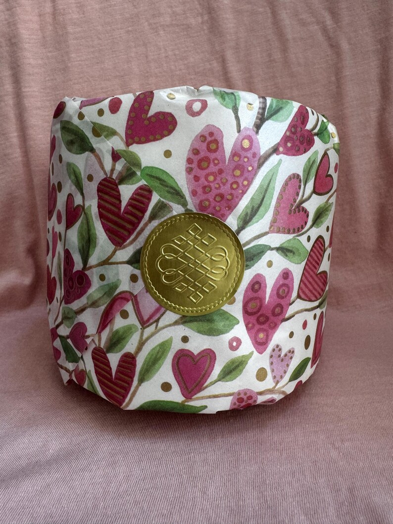 May include: A white cylinder wrapped in paper with a pink and green floral pattern featuring hearts and gold dots. A gold seal with a knot design is in the centre of the cylinder.