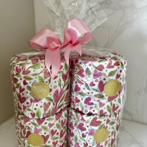 May include: Four rolls of pink and green patterned toilet paper wrapped in clear plastic with a pink satin ribbon bow. The toilet paper has a gold foil sticker with the word "Love" on it.