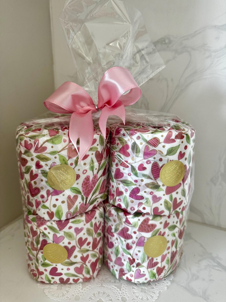 May include: Four rolls of pink and green patterned toilet paper wrapped in clear plastic with a pink satin ribbon bow. The toilet paper has a gold foil sticker with the word "Love" on it.