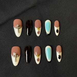 May include: Set of ten false nails with different designs.  The nails are almond shaped and have a white base with different colored tips.  Some nails have a cross design with rhinestones, others have a leopard print, and others have a gold star design.