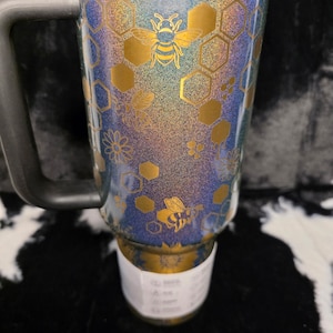 以下が含まれることがあります： 黒いハンドルと蓋が付いたトラベルマグ。金色のバンドと蜂とハニカムの模様が特徴です。マグは青、紫、金のグリッター仕上げです。「Happy Poms Creations」の文字が下部にあります。