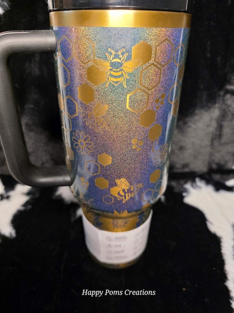 以下が含まれることがあります： 黒いハンドルと蓋が付いたトラベルマグ。金色のバンドと蜂とハニカムの模様が特徴です。マグは青、紫、金のグリッター仕上げです。「Happy Poms Creations」の文字が下部にあります。