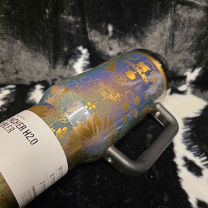 以下が含まれることがあります： 金と虹色の花柄が施されたステンレス製のタンブラー。タンブラーには黒いハンドルと「Quencher H2.0 Tumbler」の文字が入った白いラベルが付いています。タンブラーはヒマワリやその他の花柄で装飾されています。
