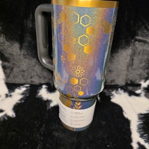 以下が含まれることがあります： 黒いハンドルと蓋が付いたキラキラ光るタンブラー。タンブラーの上部には金のバンドがあり、金色のハニカムと花の模様で装飾されています。タンブラーの底には白いラベルが付いています。「Happy Poms Creations」というテキストが下にあります。