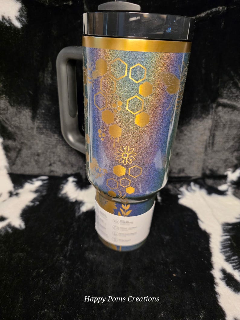 以下が含まれることがあります： 黒いハンドルと蓋が付いたキラキラ光るタンブラー。タンブラーの上部には金のバンドがあり、金色のハニカムと花の模様で装飾されています。タンブラーの底には白いラベルが付いています。「Happy Poms Creations」というテキストが下にあります。