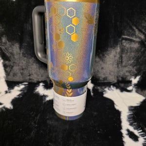 以下が含まれることがあります： 黒いハンドルと蓋が付いた、虹色のタンブラー。タンブラーは、金色のハニカムと花の模様が特徴です。上部の縁は金色です。「Happy Poms Creations」の文字が下部にあります。