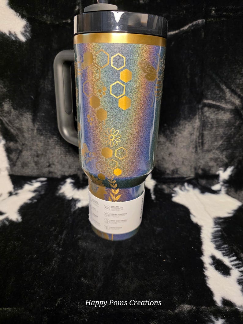 以下が含まれることがあります： 黒いハンドルと蓋が付いた、虹色のタンブラー。タンブラーは、金色のハニカムと花の模様が特徴です。上部の縁は金色です。「Happy Poms Creations」の文字が下部にあります。
