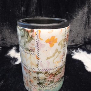 May include: A white and floral patterned can cooler with a black rim. The design features a patchwork style with various floral and ribbon motifs. The bottom section has a glittery, light blue finish. The text "Happy Poms Creations" is at the bottom.