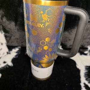 以下が含まれることがあります： 黒いハンドルと蓋が付いたスタンレーのトラベルマグ。マグには金のバンドがあり、金の蜂、花、ハニカム模様で装飾されています。マグは青、紫、金のグリッターデザインです。