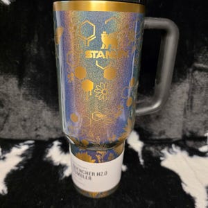 以下が含まれることがあります： 黒いハンドルと蓋が付いたスタンレーQuencher H2.0タンブラー。タンブラーは、スタンレーのロゴが入った金と青の花柄のデザインが特徴です。タンブラーの底には「Quencher H2.0 Tumbler」と書かれた白いラベルが付いています。