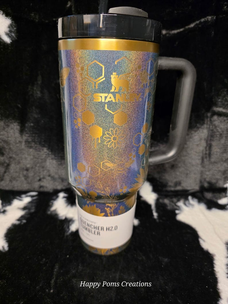 以下が含まれることがあります： 黒いハンドルと蓋が付いたスタンレーQuencher H2.0タンブラー。タンブラーは、スタンレーのロゴが入った金と青の花柄のデザインが特徴です。タンブラーの底には「Quencher H2.0 Tumbler」と書かれた白いラベルが付いています。