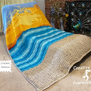 May include: A crocheted blanket with a beach scene design. The blanket features a blue sky, yellow sun, and blue waves. The blanket is draped over a chair and has the text "Creative Art Expressions" on the bottom right corner.