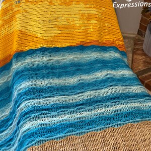 May include: A crocheted blanket with a beach scene. The blanket is made of different colored yarn to create the look of a beach with sand, waves, and sky. The blanket is folded in half and the top half is folded over the bottom half.