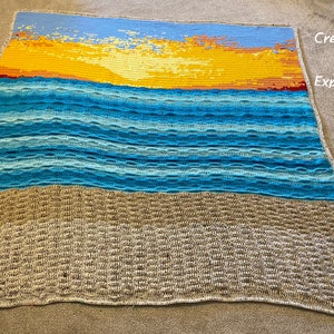 May include: A crocheted blanket with a sunset over the ocean scene. The blanket is made with different shades of blue, yellow, and brown yarn. The blanket is on a gray surface. Creative Art Expressions is written in the top right corner.