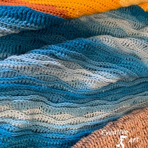 May include: A close-up of a crocheted blanket with a blue and white ombre pattern. The blanket is folded in a wave-like pattern, with a yellow and orange crocheted blanket visible in the background. The text "Creative Art Expressions" is visible in the bottom right corner of the image.