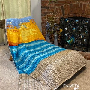May include: A crocheted blanket with a beach scene. The blanket features a sunset over the ocean with blue and white waves. The blanket is draped over a chair in a living room.