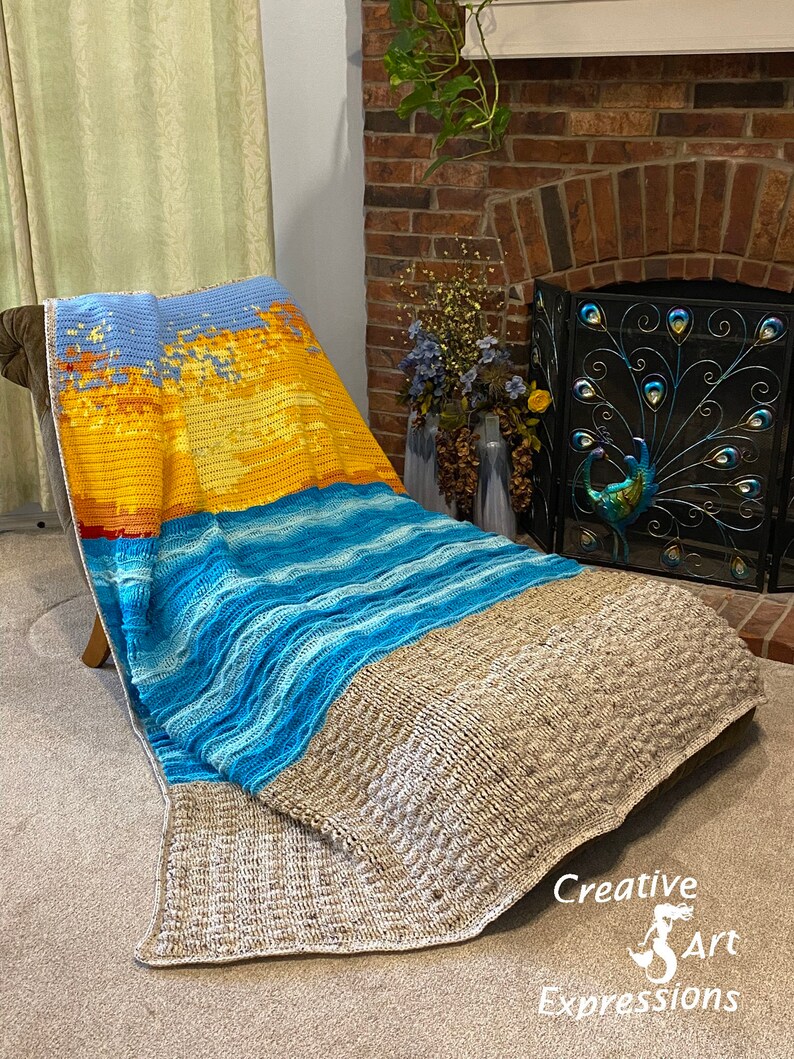 May include: A crocheted blanket with a beach scene. The blanket features a sunset over the ocean with blue and white waves. The blanket is draped over a chair in a living room.