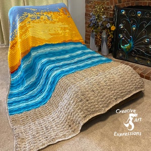 May include: A crocheted blanket with a beach scene. The blanket is made with different shades of blue, yellow, and brown yarn. The blanket is draped over a chair. The text "Creative Art Expressions" is visible in the lower right corner of the image.