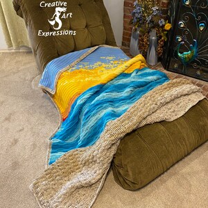 May include: A crocheted blanket with a blue, yellow, and beige color scheme draped over a brown armchair. The blanket has a wave pattern and is made with a variety of textures. The armchair has a tufted back and is in a room with a fireplace.