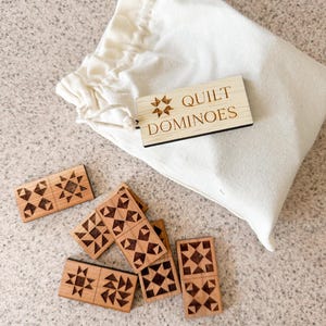 May include: A set of wooden dominoes with a quilt pattern design, scattered on a surface next to a drawstring bag. The dominoes are light brown with dark brown etched patterns. The bag is off-white, and a wooden tag reads "Quilt Dominoes."