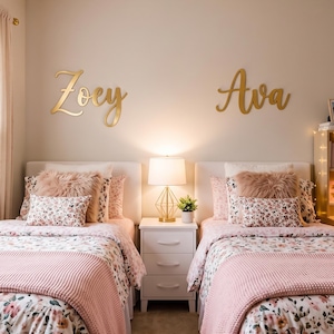 Pode incluir: Duas camas de solteiro com cabeceiras brancas, roupa de cama com estampa floral e mantas de tricô rosa. Uma mesa de cabeceira branca de três gavetas fica entre as camas com uma luminária dourada. Os nomes Zoey e Ava estão em letras douradas.