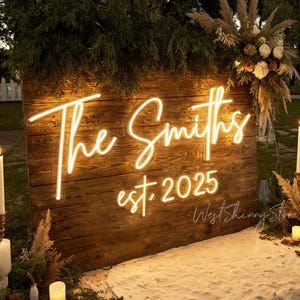 May include: A wooden sign with the words "The Smiths" in a glowing neon script, with "est. 2025" below. The sign is decorated with greenery and flowers, perfect for a wedding or anniversary celebration.