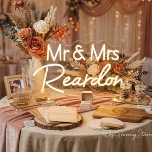May include: A wedding reception table is decorated with floral arrangements, a glowing "Mr & Mrs Reardon" sign, and various wedding-themed items. The table includes a ring box, place cards, and a wooden cutting board.
