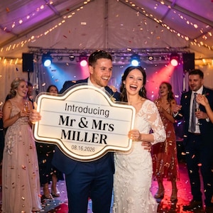 May include: A newly married couple smiles, holding a light-up sign that reads "Introducing Mr & Mrs Miller" with the date 08.16.2026. The wedding reception is filled with guests, confetti, and decorative lights.