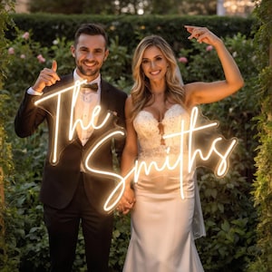 May include: A couple poses with a neon sign that reads "The Smiths" in cursive script. The sign is illuminated with a warm, white light. The woman wears a white wedding dress, and the man wears a black tuxedo. They are standing in a garden setting.