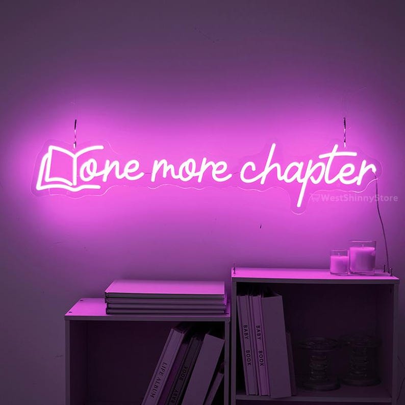 May include: A pink neon sign with the text "One more chapter" and a stylized book icon. The sign is hanging on a wall with a bookshelf below it.