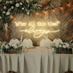 May include: A wedding reception table setting with a white tablecloth and greenery centerpieces. A neon sign with the text "after all this time? always" is displayed on the wall behind the table.