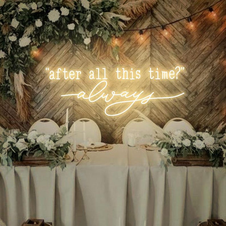 May include: A wedding reception table setting with a white tablecloth and greenery centerpieces. A neon sign with the text "after all this time? always" is displayed on the wall behind the table.