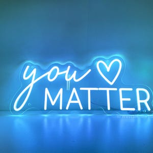May include: White neon sign with the words "you" and a heart symbol, followed by the word "matter". The sign is lit up and glowing against a blue background.