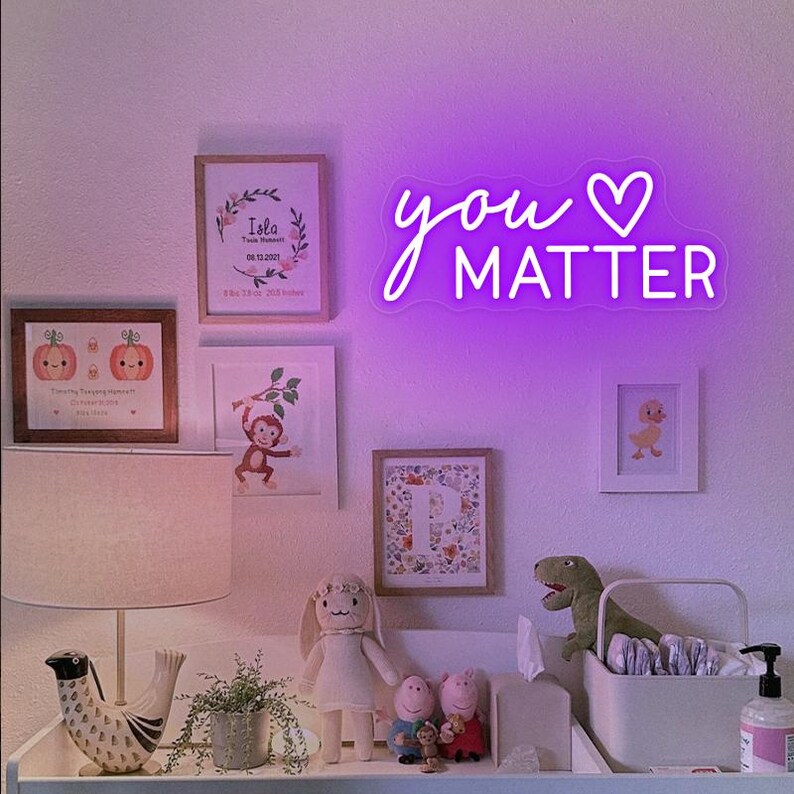 May include: A purple neon sign with the text "you &hearts; MATTER" hanging on a white wall. The sign is surrounded by framed pictures and other decorations.