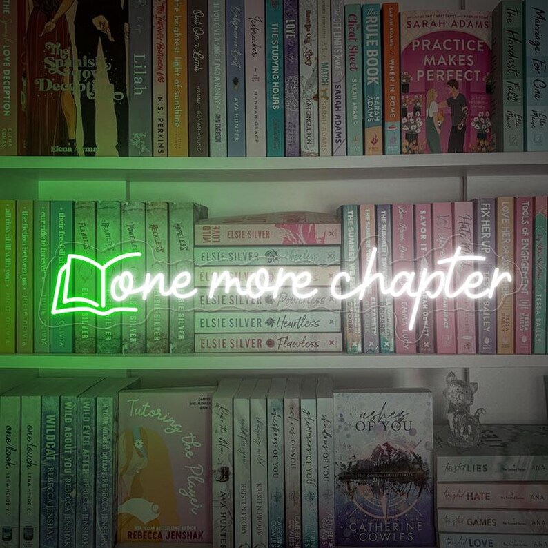 May include: A green neon sign with a book icon and the text "One more chapter" displayed on a bookshelf with a variety of books.