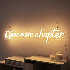 May include: White neon sign with the words "One more chapter" in a cursive font. The sign is hanging on a white wall above a shelf with books.