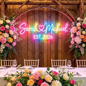 Puede incluir: Una mesa de recepción de bodas está decorada con un arreglo floral de rosas rosas, amarillas y blancas. Un letrero de neón muestra "Sarah & Michael EST. 2026" sobre la mesa. El fondo es una pared de madera.