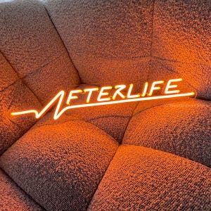 May include: A neon sign with the word "AFTERLIFE" in a yellow-orange colour. The sign is on a brown fabric surface.