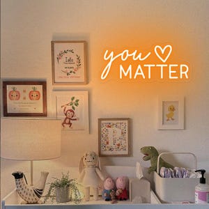 May include: A nursery room with a white wall and a neon sign that says "you matter" with a heart. There are several framed pictures on the wall, including one with a monkey, one with a duck, and one with a pumpkin. There is a lamp on a table with a bird figurine, a potted plant, and a stuffed animal.