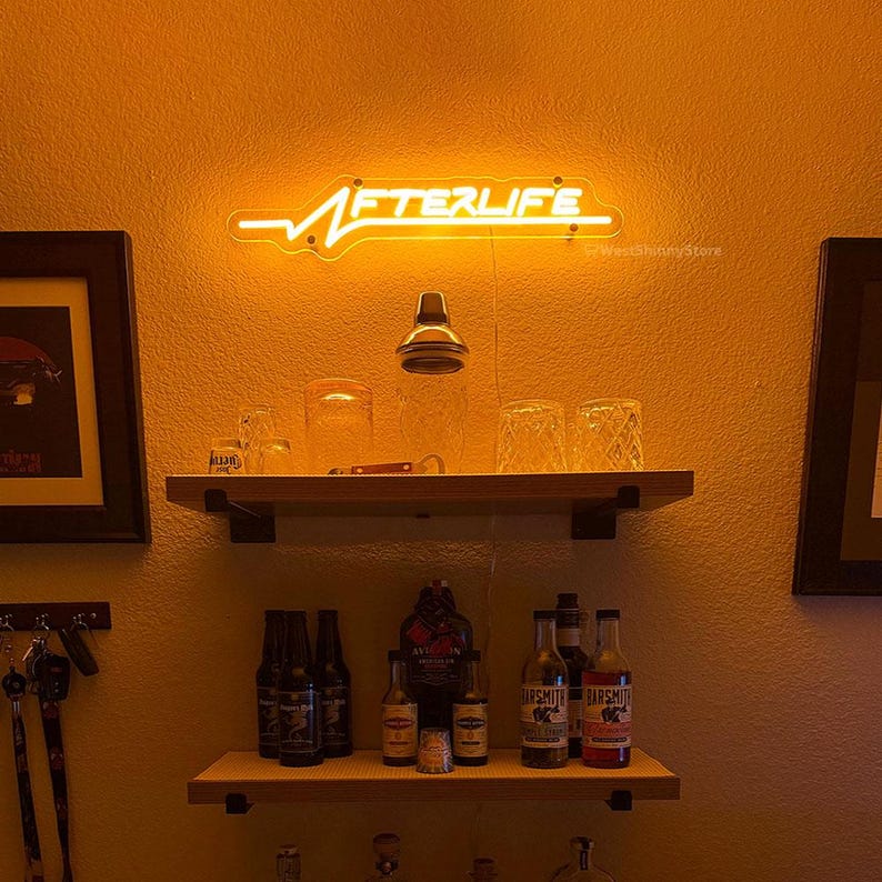 Op de afbeelding: Een neonbord met het woord "AFTERLIFE" in witte letters op een gele achtergrond. Het bord is gemonteerd aan een muur boven een plank met verschillende flessen en glazen.
