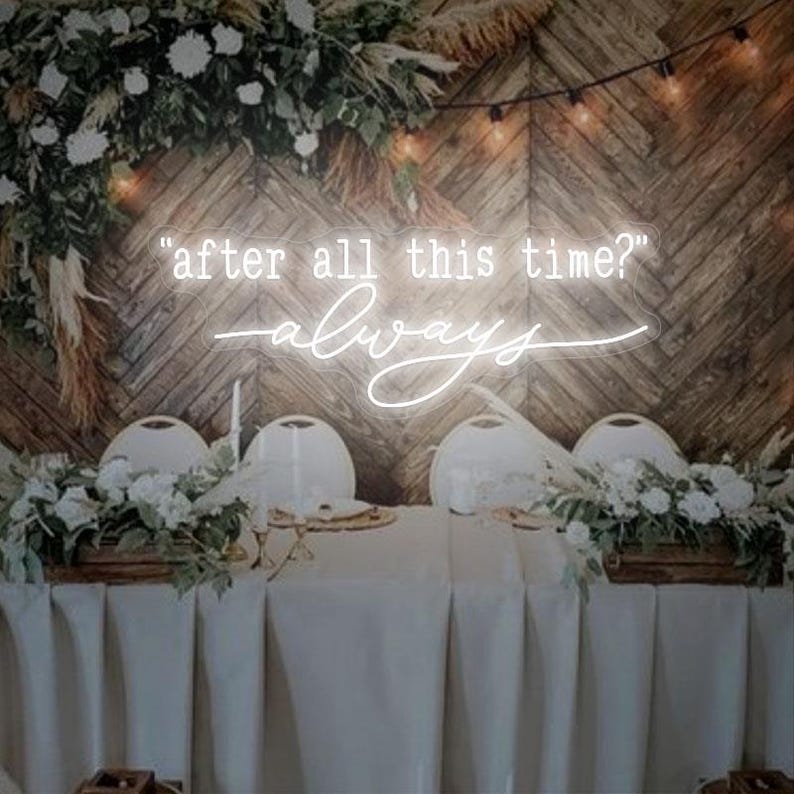 May include: A white neon sign with the quote "after all this time? always" on a wooden wall backdrop. The sign is displayed above a table set for a wedding reception with white linens and floral centerpieces.