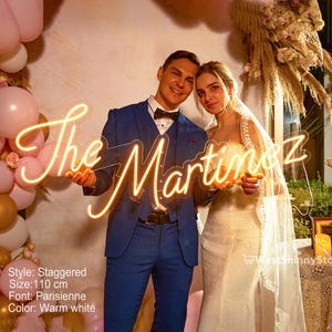 May include: A warm white neon sign that reads "The Martinez" in a cursive font. The sign is held by a couple, with the man in a blue suit and the woman in a wedding dress. The sign is 110 cm in size.
